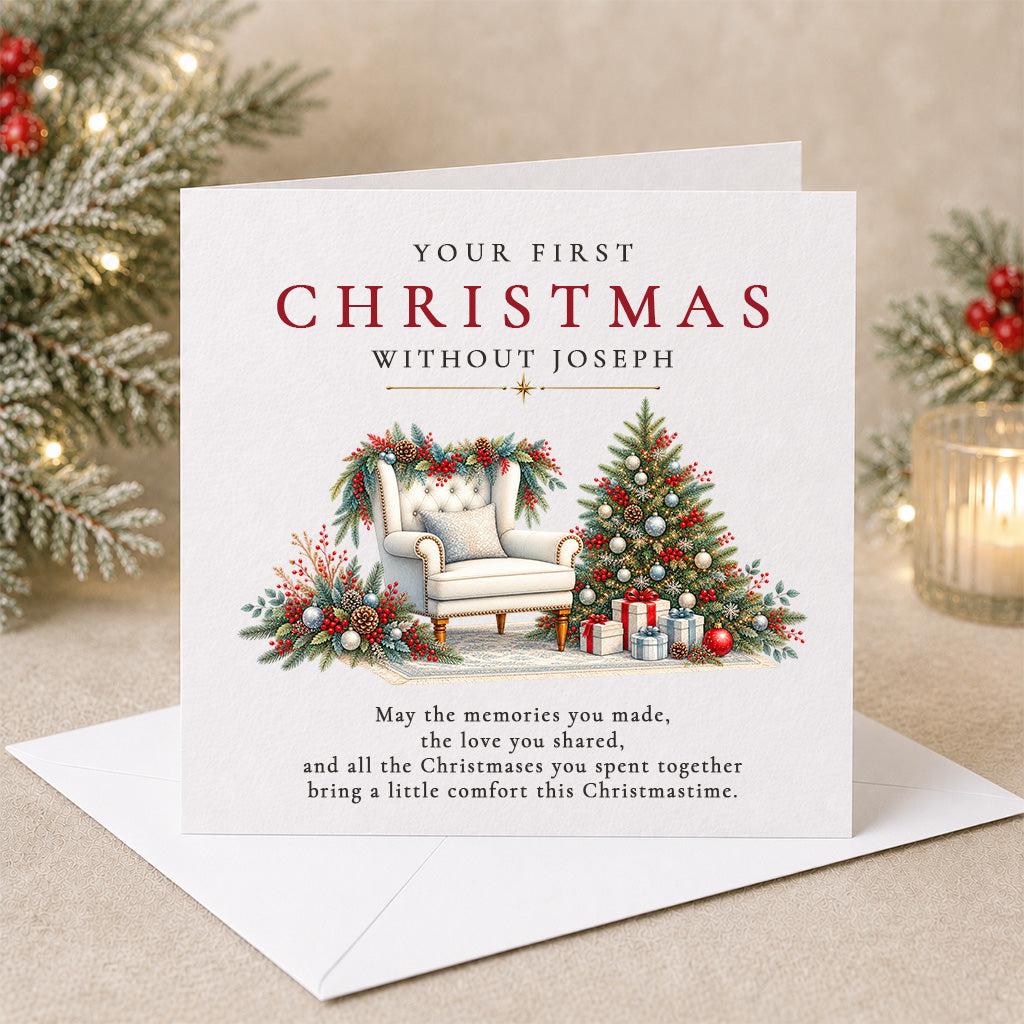 A personalized Christmas greeting card with an envelope, displayed on a beige surface with festive blurred holiday decorations in the background. The card reads 'YOUR FIRST CHRISTMAS WITHOUT JOSEPH' and features an illustration of a cozy armchair, a decorated Christmas tree, and wrapped gifts.