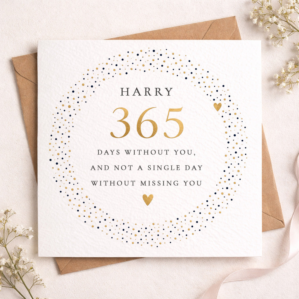A square white greeting card with a circular dotted wreath design in black and gold, featuring the text 'HARRY 365 DAYS WITHOUT YOU, AND NOT A SINGLE DAY WITHOUT MISSING YOU,' styled on top of a brown envelope with dried flowers and a pale pink ribbon.