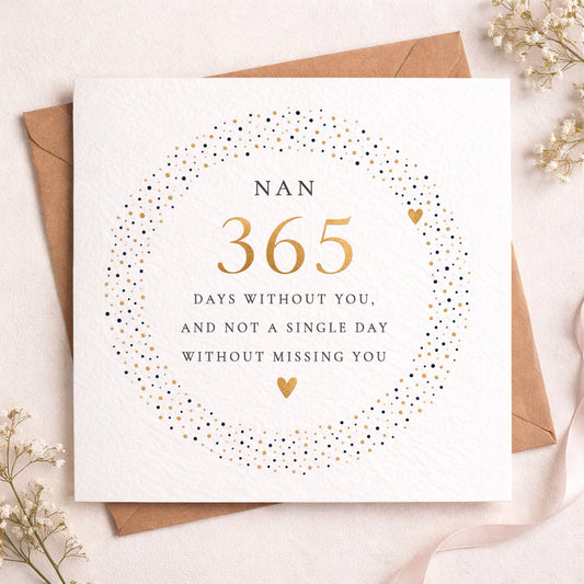 A square white greeting card with a circular dotted border in black and gold, reading 'NAN 365 DAYS WITHOUT YOU, AND NOT A SINGLE DAY WITHOUT MISSING YOU,' styled on top of a brown envelope with small dried flowers around it.