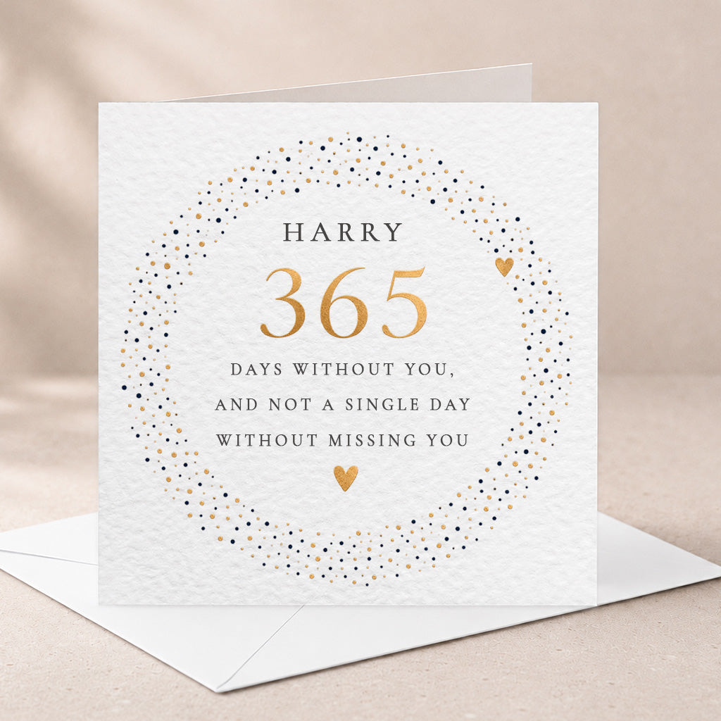 A square white greeting card with a textured paper finish, featuring a circular gold and black dot wreath design and the text 'HARRY 365 DAYS WITHOUT YOU, AND NOT A SINGLE DAY WITHOUT MISSING YOU.' It is shown standing upright with an envelope behind it on a soft beige surface.
