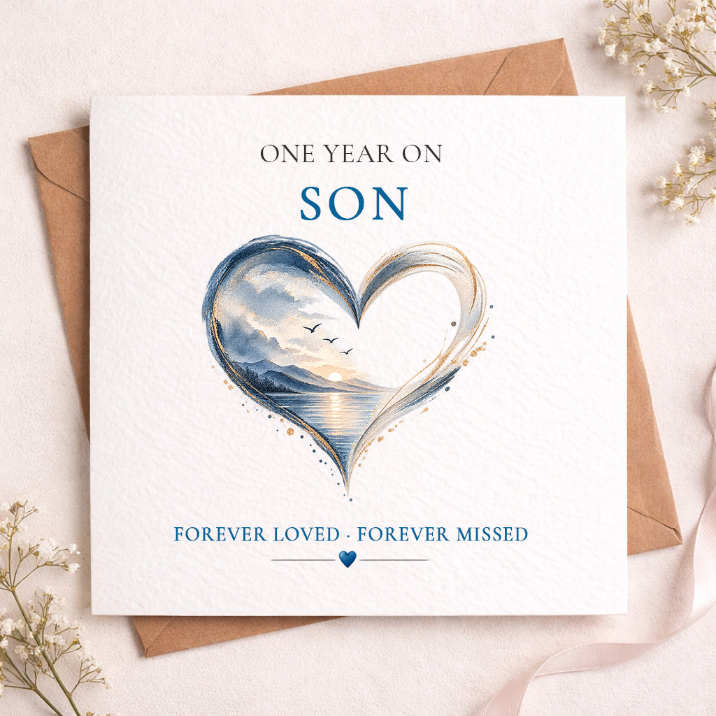 A square white memorial card with a watercolor-style heart illustration in blue, gray, and warm golden tones, placed on top of a brown envelope. The card reads 'ONE YEAR ON SON' and 'FOREVER LOVED • FOREVER MISSED' with a small blue heart at the bottom.