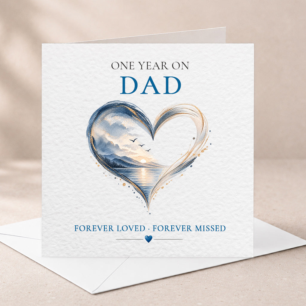 A square white greeting card with an envelope behind it, printed with the text 'ONE YEAR ON DAD' and 'FOREVER LOVED - FOREVER MISSED' around a large heart-shaped scenic illustration in blue, gray, and warm golden tones.