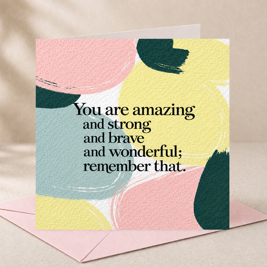 A square greeting card with abstract pastel brushstrokes in pink, yellow, and light blue, overlaid with black text reading, 'You are amazing and strong and brave and wonderful; remember that.' A pink envelope lies behind it on a soft beige surface.