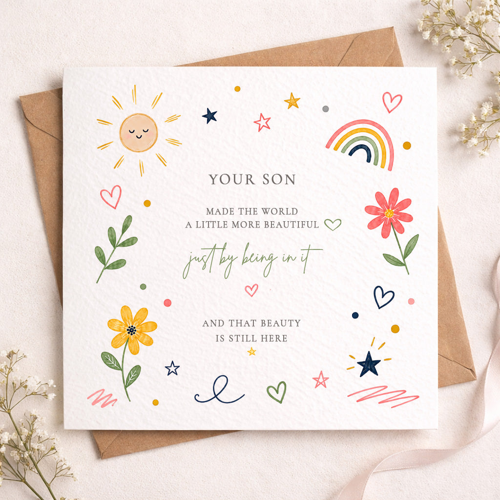 A square white greeting card with colorful hand-drawn illustrations and a brown envelope behind it, styled on a light beige surface with small dried flowers and ribbon.