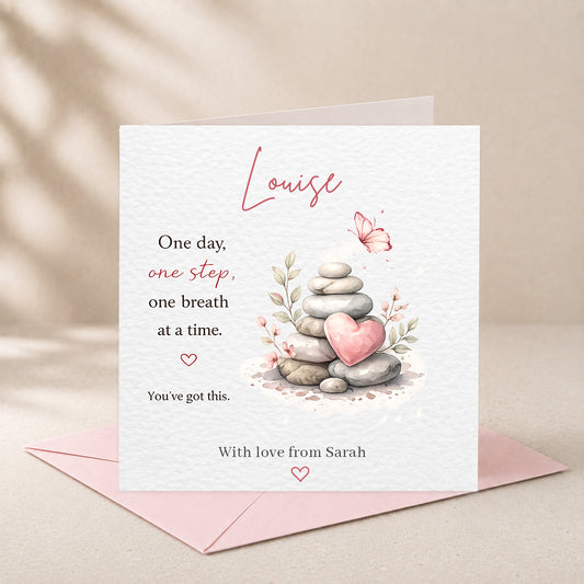 A square white greeting card with a pink envelope behind it, featuring a watercolor-style stack of smooth stones with a pink heart, a butterfly, and printed message text. The card is shown standing upright on a soft beige surface with a shadow pattern in the background.