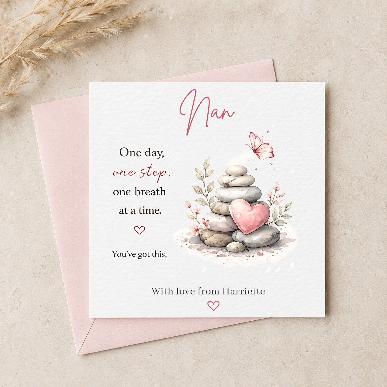 A square greeting card for a Nan, placed on top of a light pink envelope. The card has a white background with a watercolor illustration of stacked gray stones, a pink heart, and a butterfly, alongside the quote, 'One day, one step, one breath at a time.