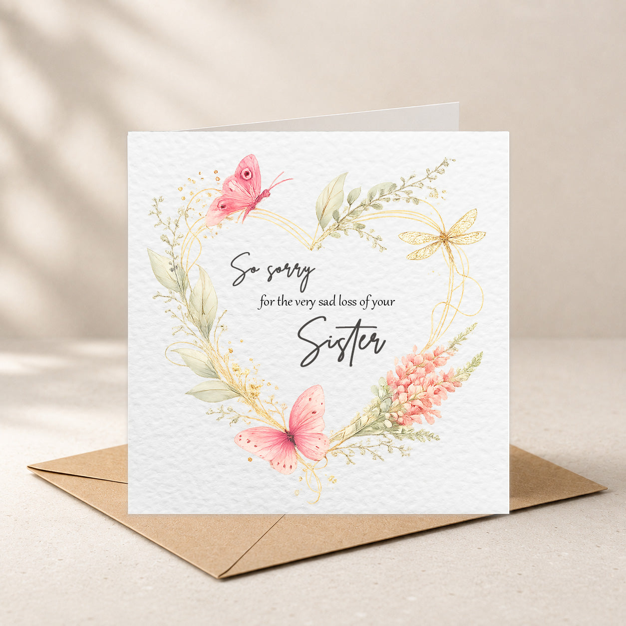 A square white greeting card with a floral wreath and butterflies around the text 'So sorry for the very sad loss of your Sister,' shown standing on top of a brown envelope against a soft beige background.