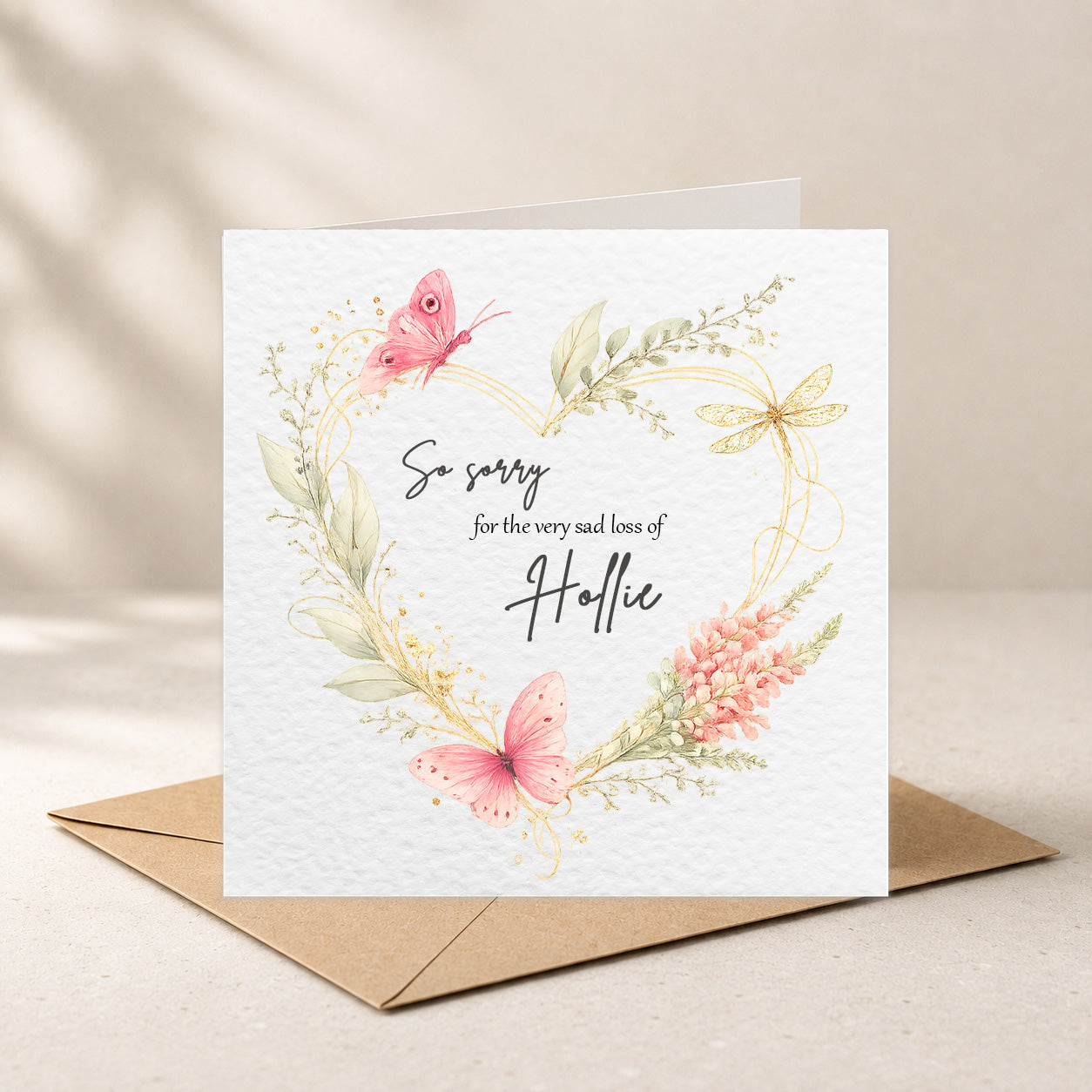 A square white greeting card with a watercolor floral wreath and butterflies, reading 'So sorry for the very sad loss of Helle,' displayed with a brown envelope on a light beige surface.