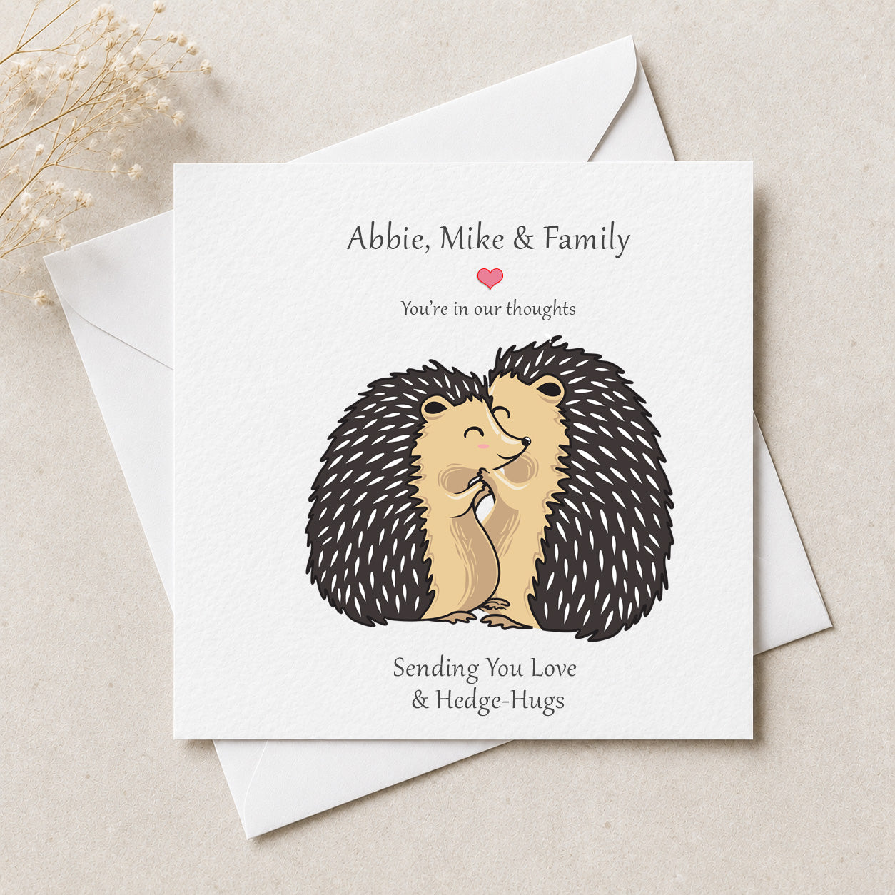 A square greeting card with a white background, featuring an illustration of two hugging hedgehogs and the text 'Abbie, Mike & Family You're in our thoughts Sending You Love & Hedge-Hugs'. The card is styled on top of white envelopes on a light beige textured surface with dried flowers in the corner.
