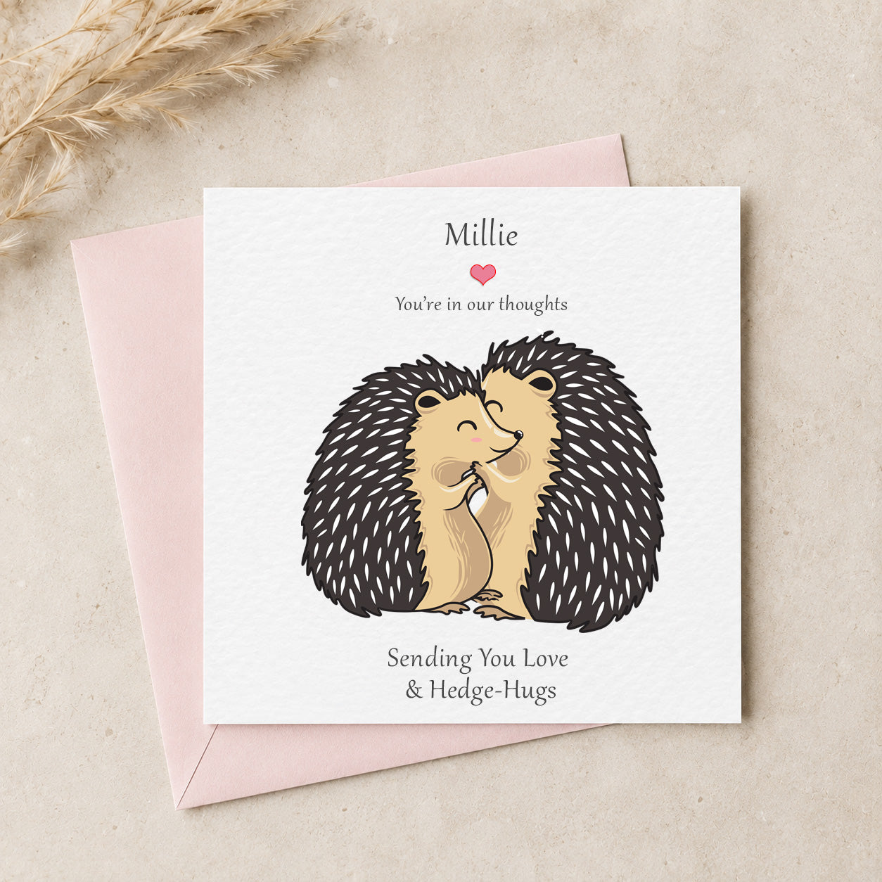 A personalized greeting card featuring an illustration of two hugging hedgehogs, with the text 'Millie You're in our thoughts' and 'Sending You Love & Hedge-Hugs'. The card is shown with a light pink envelope on a neutral beige textured background with dried grass in the corner.