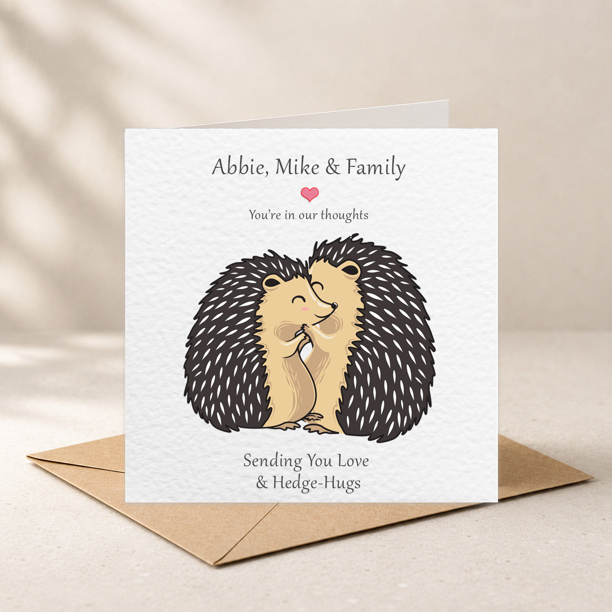 A square white greeting card with a cute hedgehog illustration and the text 'Abbie, Mike & Family You're in our thoughts Sending You Love & Hedge-Hugs', shown standing upright on a brown envelope against a warm beige background.
