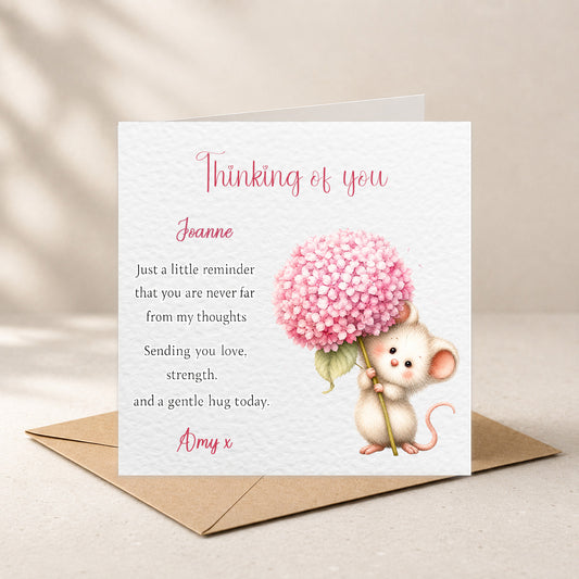A square greeting card with a white textured background, featuring a pink hydrangea illustration and a small cartoon mouse holding the stem. The card reads 'Thinking of you' and includes a message addressed to 'Joanne' from 'Amy x'.