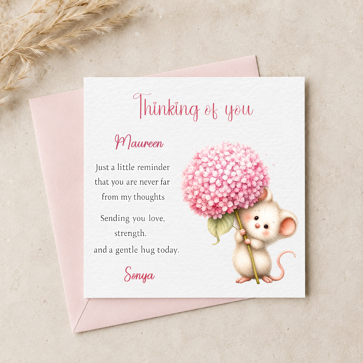 A square white greeting card with a pink envelope behind it, styled on a light beige surface with dried grass. The card reads 'Thinking of you' and includes a pink watercolor mouse holding a pink flower.