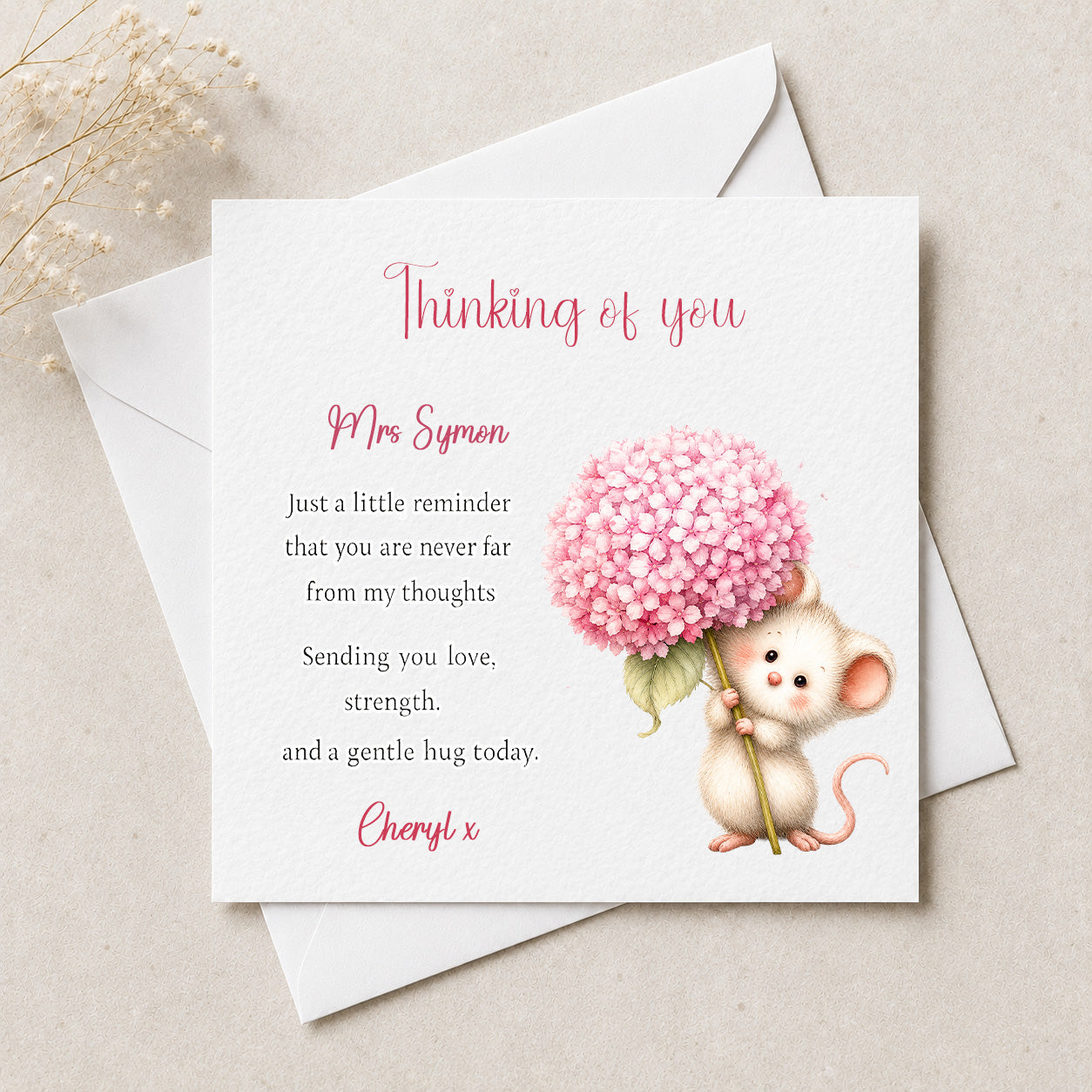 A square white greeting card with a watercolor illustration of a small mouse holding a pink flower, placed on top of white envelopes on a light beige surface. The card reads 'Thinking of you Mrs.