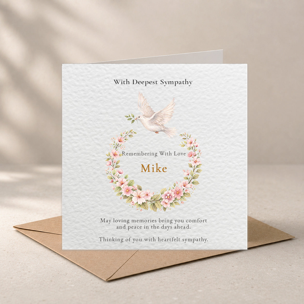 A sympathy greeting card with a floral wreath illustration and the name 'Mike' printed on it, shown standing upright with a brown envelope behind it on a neutral beige surface.