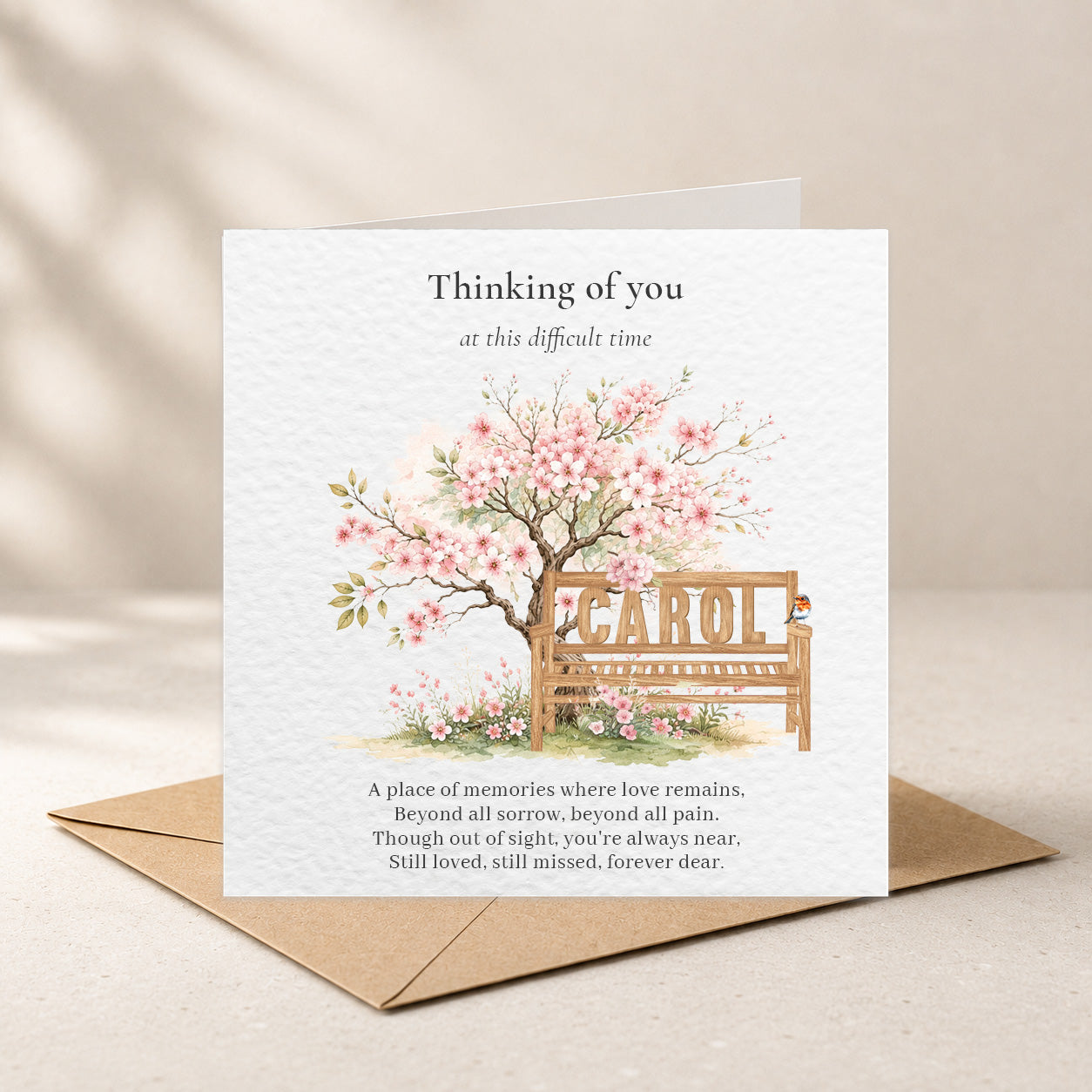 A square sympathy card with a kraft envelope, featuring a watercolor illustration of a cherry blossom tree and bench with the name 'CAROL' on the bench. The card reads 'Thinking of you at this difficult time' and includes a memorial poem at the bottom.