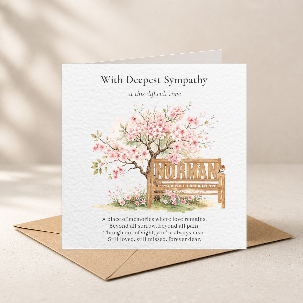 A sympathy greeting card with a kraft envelope, featuring a watercolor illustration of a cherry blossom tree and a wooden bench with the name 'NURMAN' on it, set against a warm beige background.