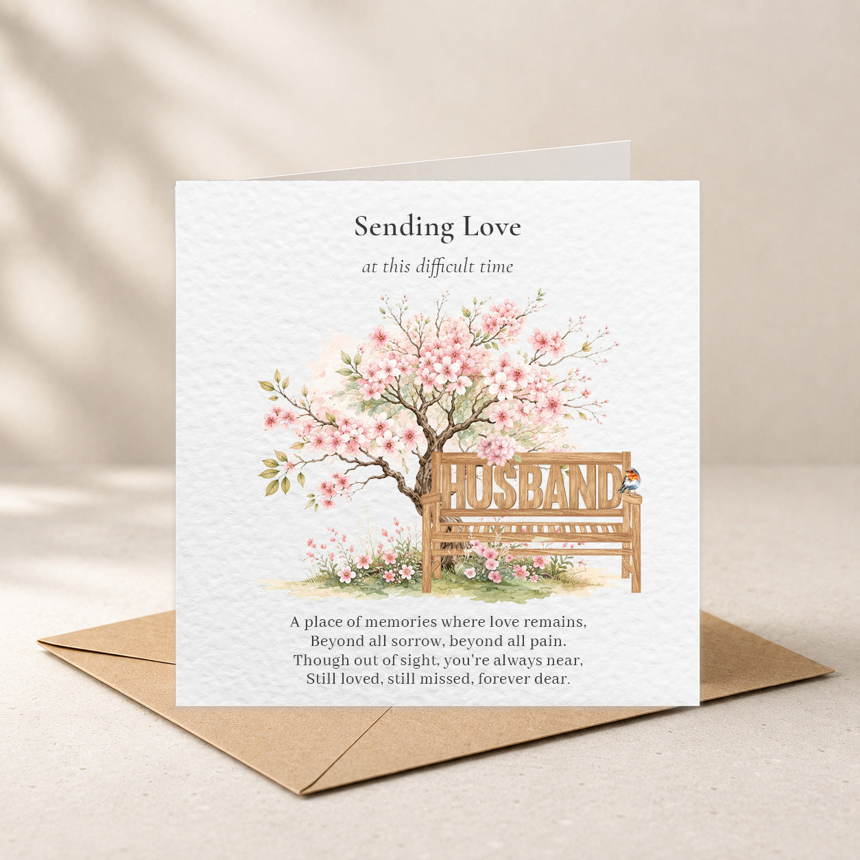 A square sympathy card with a kraft envelope, featuring a watercolor illustration of a blossoming tree and a wooden bench with the word “HUSBAND” on it. The card reads “Sending Love at this difficult time” and includes a memorial-style poem, shown on a warm beige background.