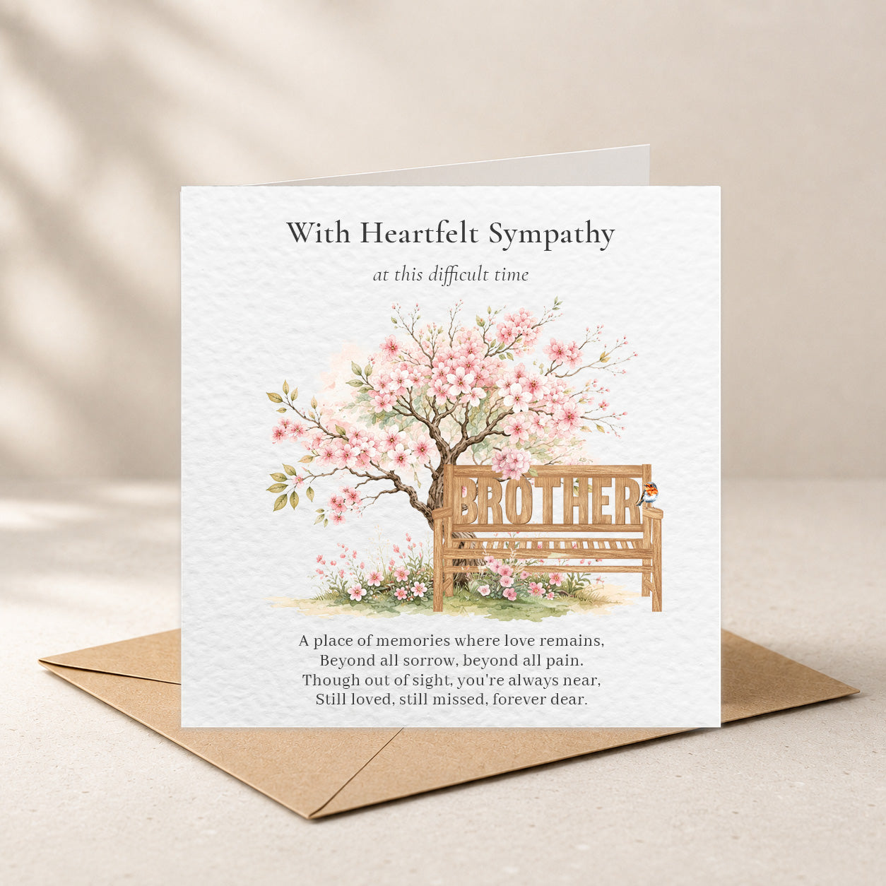A square sympathy greeting card with a kraft envelope, featuring a watercolor illustration of a cherry blossom tree and bench with the word 'BROTHER' on the seat, photographed on a warm beige surface.