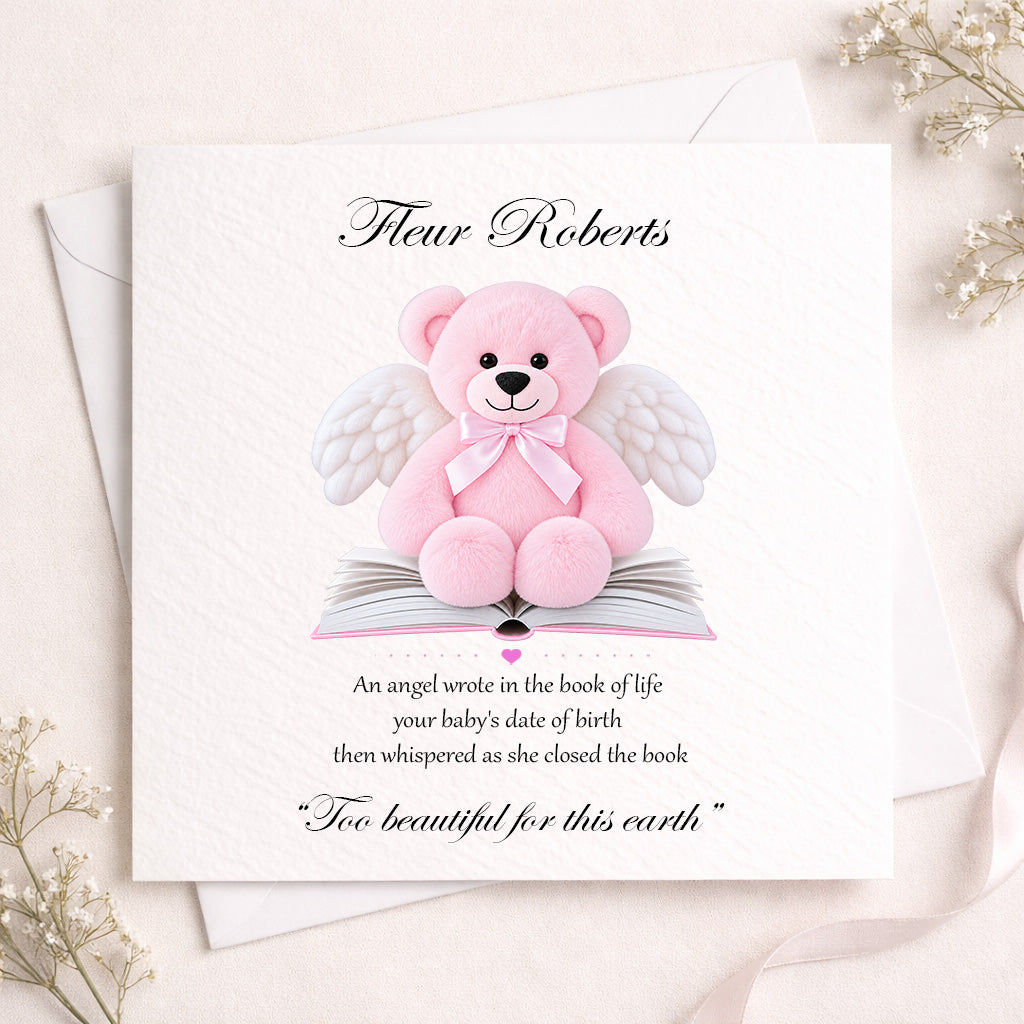 A square white memorial card featuring a pink teddy bear with white angel wings sitting on an open book, with the personalized name 'Fleur Roberts' at the top and a memorial quote below. The card is styled on a light beige surface with white envelopes and delicate dried flowers.