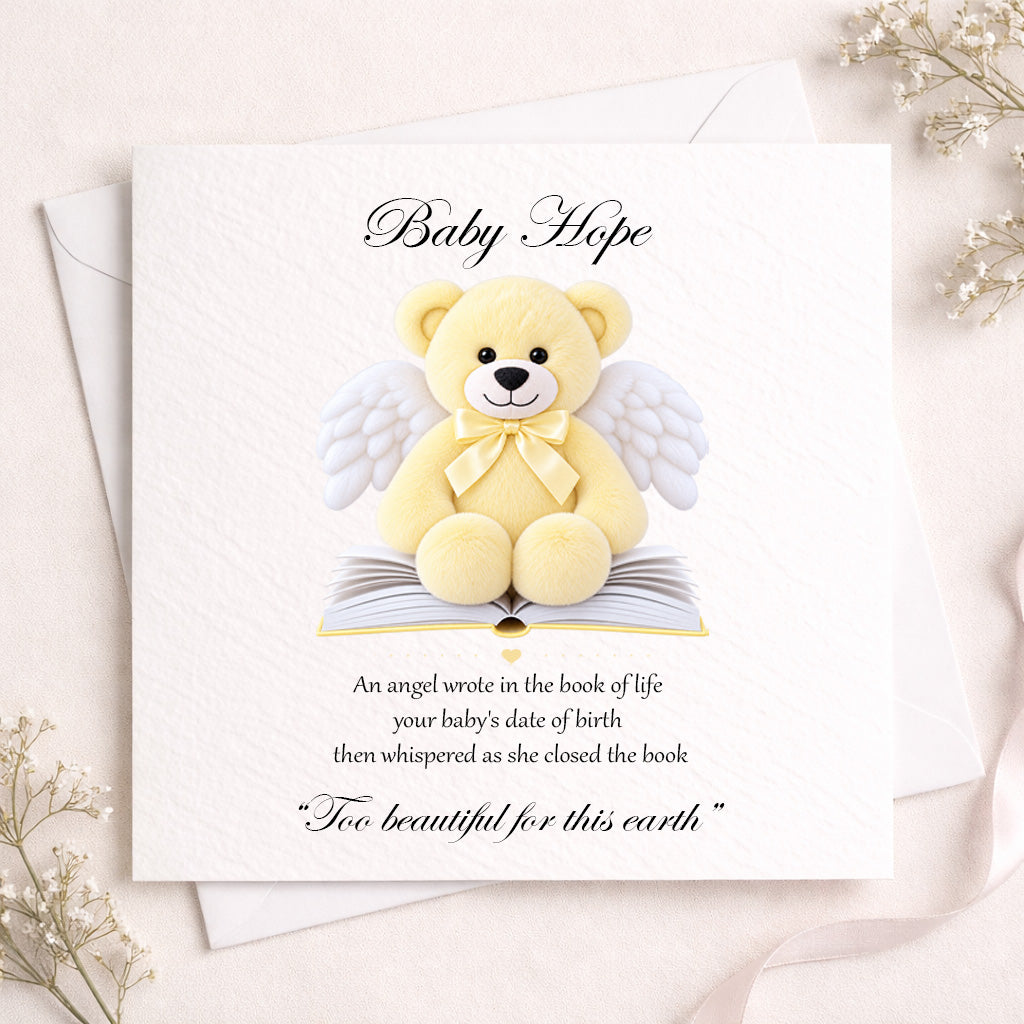 A square white baby memorial card featuring a yellow teddy bear with white angel wings sitting on an open book, with the text 'Baby Hope' and a remembrance quote. The card is styled on a light beige surface with white envelopes and delicate dried flowers.
