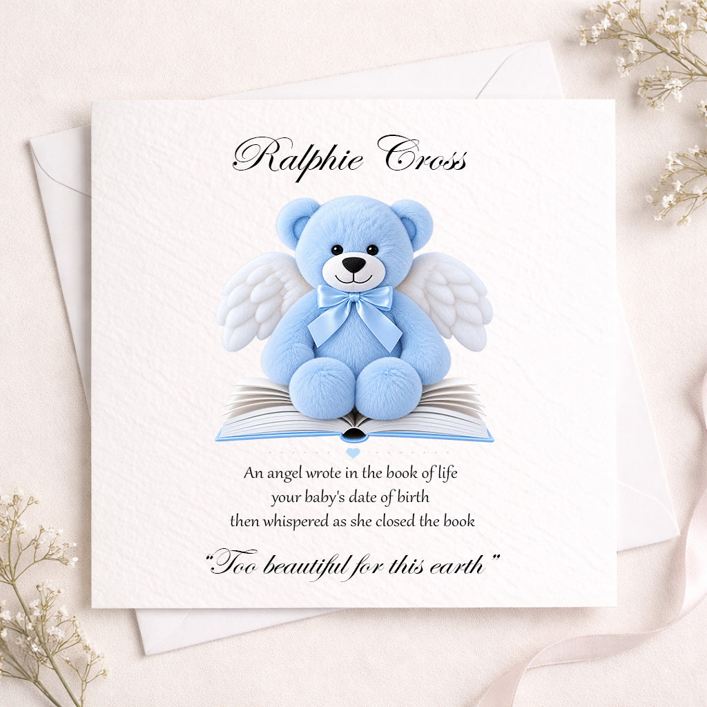 A square white memorial card featuring a blue angel teddy bear sitting on an open book, with the personalized name 'Ralphie Cross' at the top and a memorial quote below. The card is styled on top of white envelopes with delicate dried flowers in the corners.