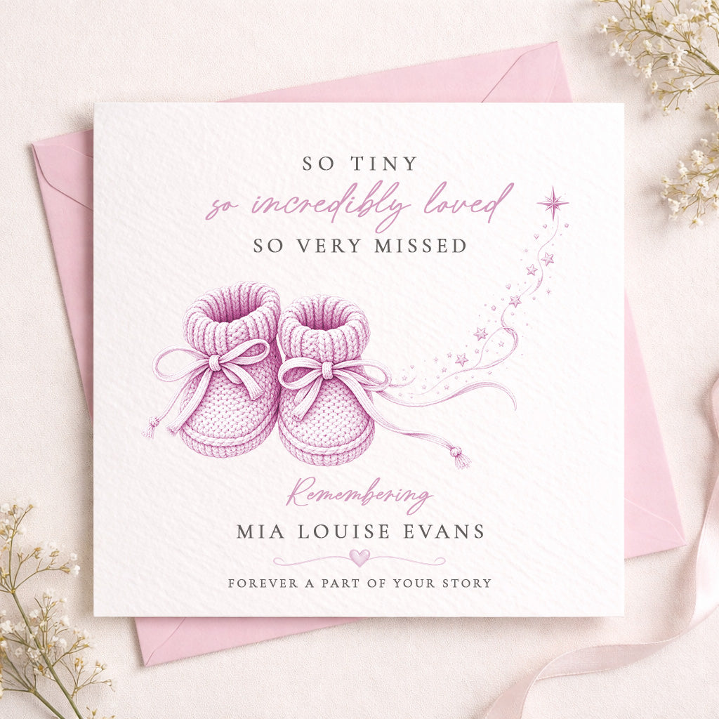 A square white memorial card with pink and dark gray text and an illustration of pink baby booties, shown with a pink envelope on a light beige surface with small white flowers around the edges.