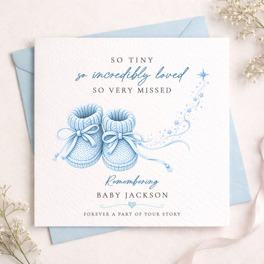 A square baby remembrance card with a white textured background, featuring a light blue illustration of baby booties and the text 'SO TINY so incredibly loved SO VERY MISSED' and 'Remembering BABY JACKSON'. The card is layered over a light blue envelope on a soft beige surface with delicate dried flowers around the edges.