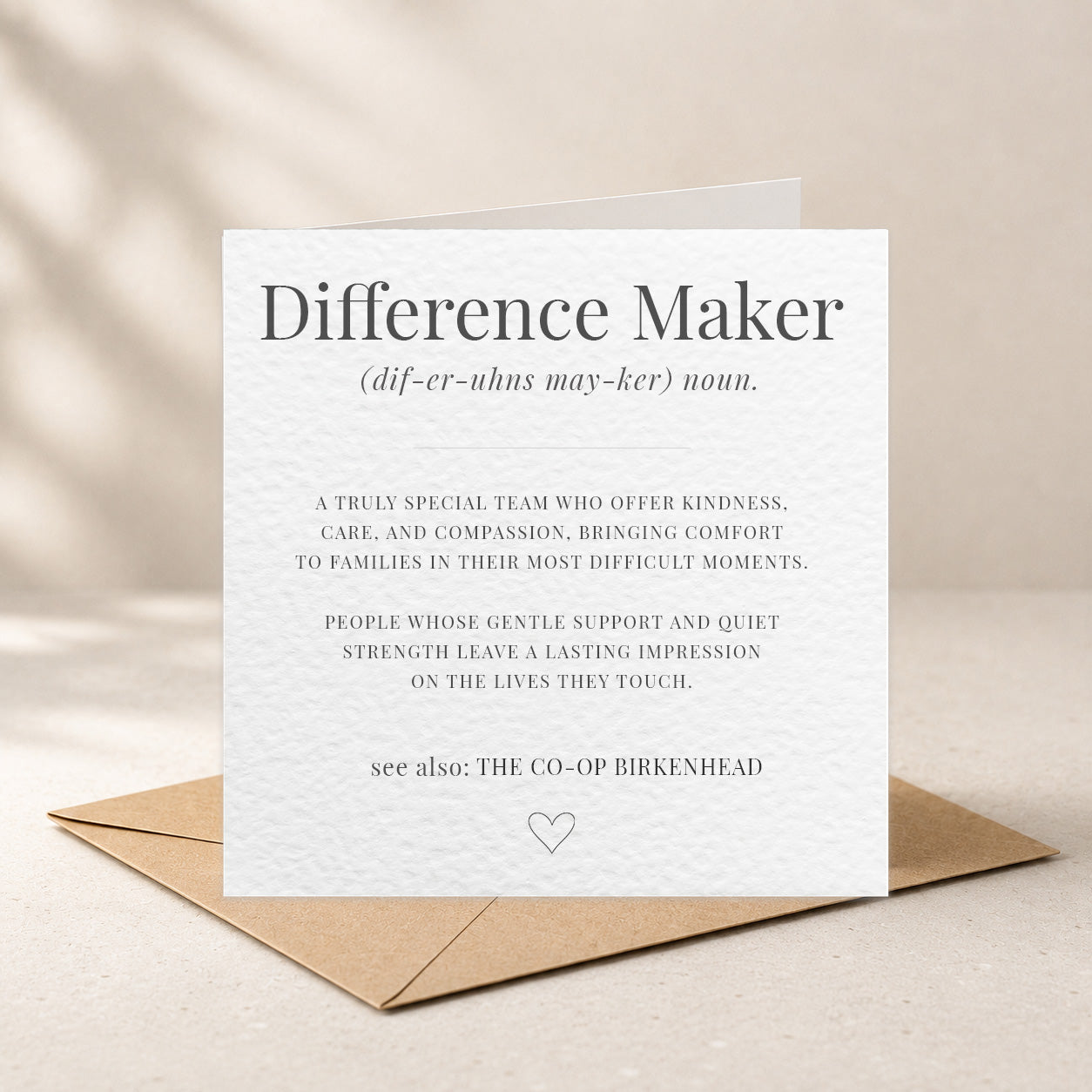 A white greeting card with black printed text reading 'Difference Maker' and a small heart at the bottom, placed on top of a brown kraft envelope against a warm beige background.