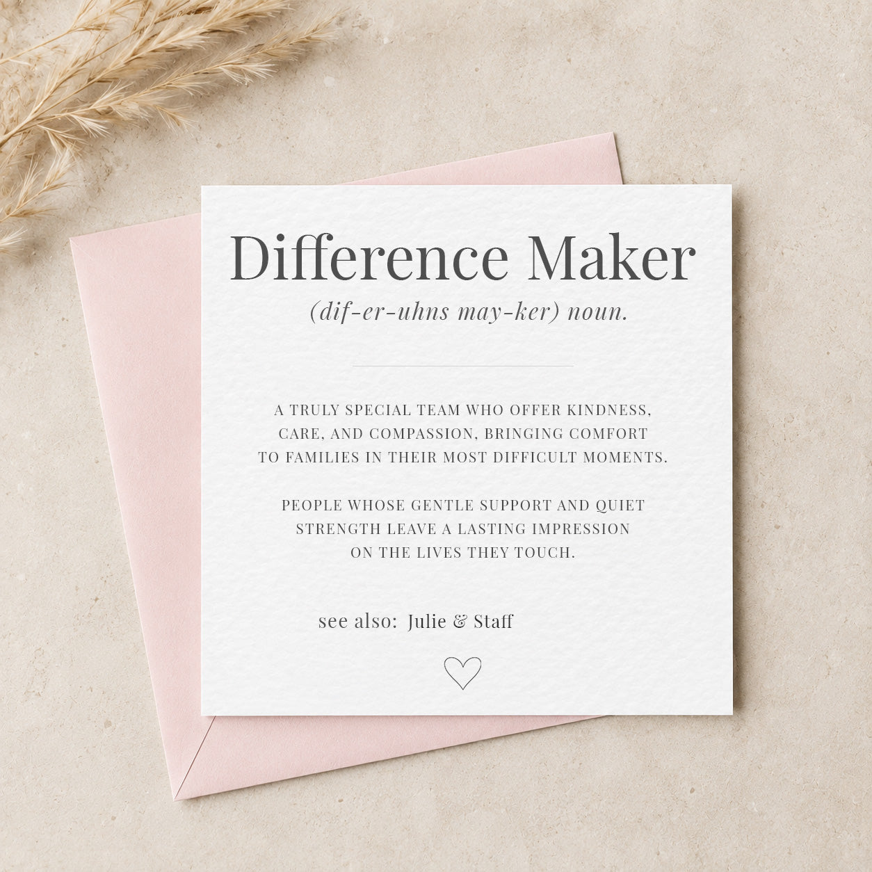 A square white greeting card with the title 'Difference Maker' in dark gray serif text, layered over a pale pink envelope on a light beige surface with dried wheat stems in the corner.