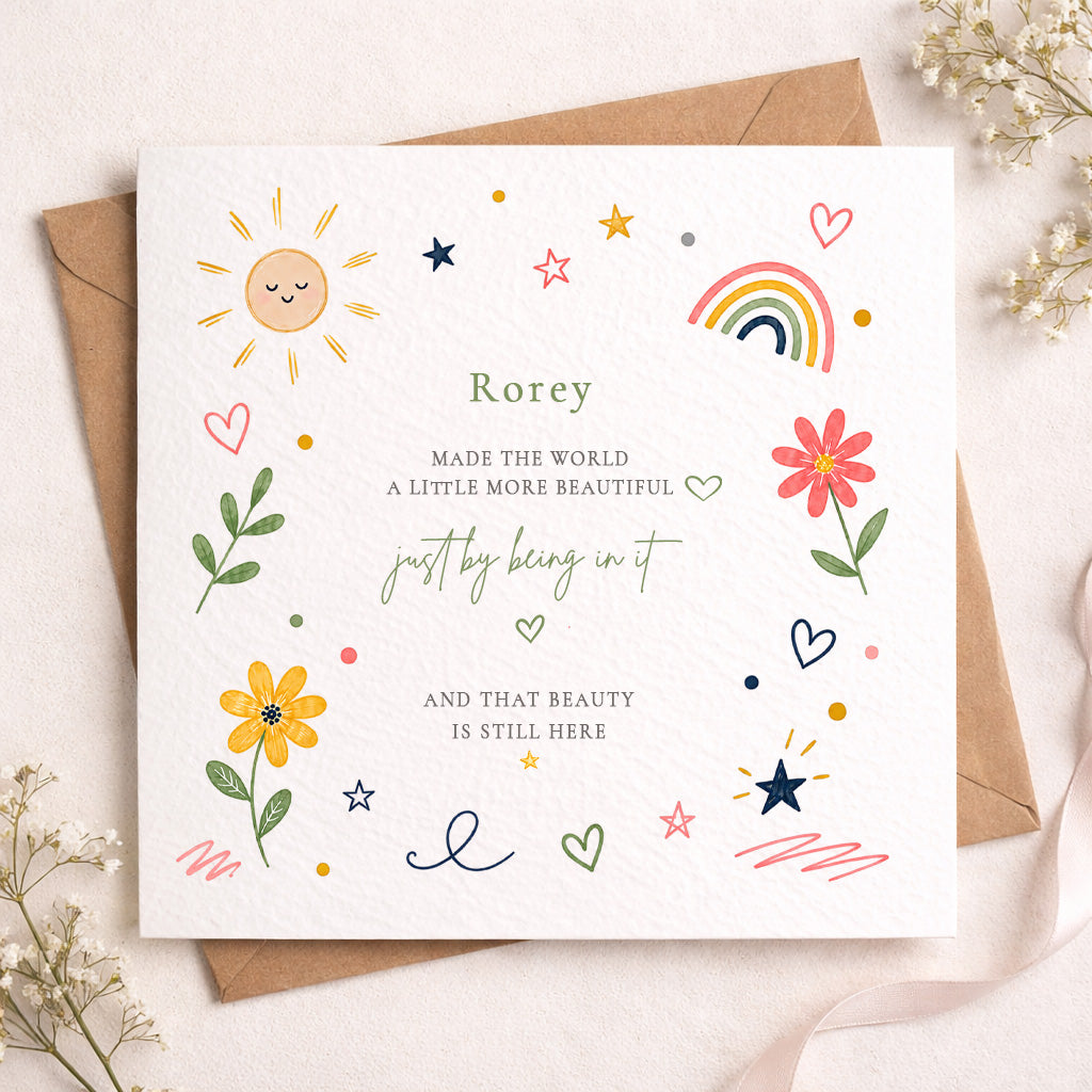 A square white greeting card with a kraft envelope behind it, decorated with colorful hand-drawn illustrations including a sun, rainbow, flowers, hearts, stars, and personalized text reading 'Rorey'.
