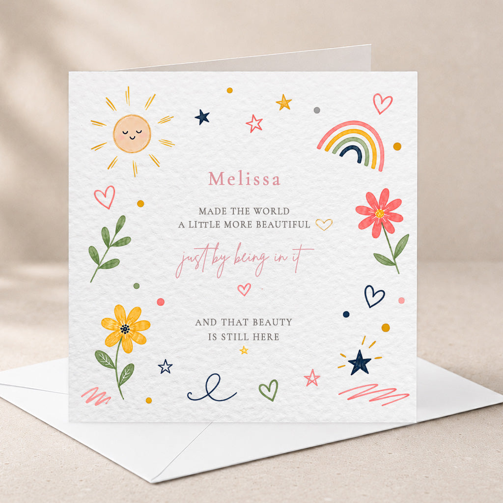A square white greeting card with a handwritten-style message and colorful doodles, shown standing on an envelope against a soft beige background. The card features a sun, rainbow, flowers, hearts, stars, and the name Melissa.