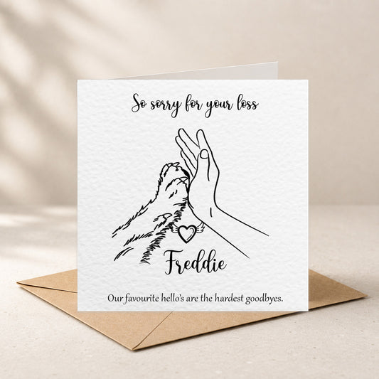 A square white sympathy card with a black line drawing of a hand holding a dog's leash, with the text 'So sorry for your loss' and 'Our favourite hello's are the hardest goodies.' The card is shown standing upright on a brown envelope against a warm beige background.