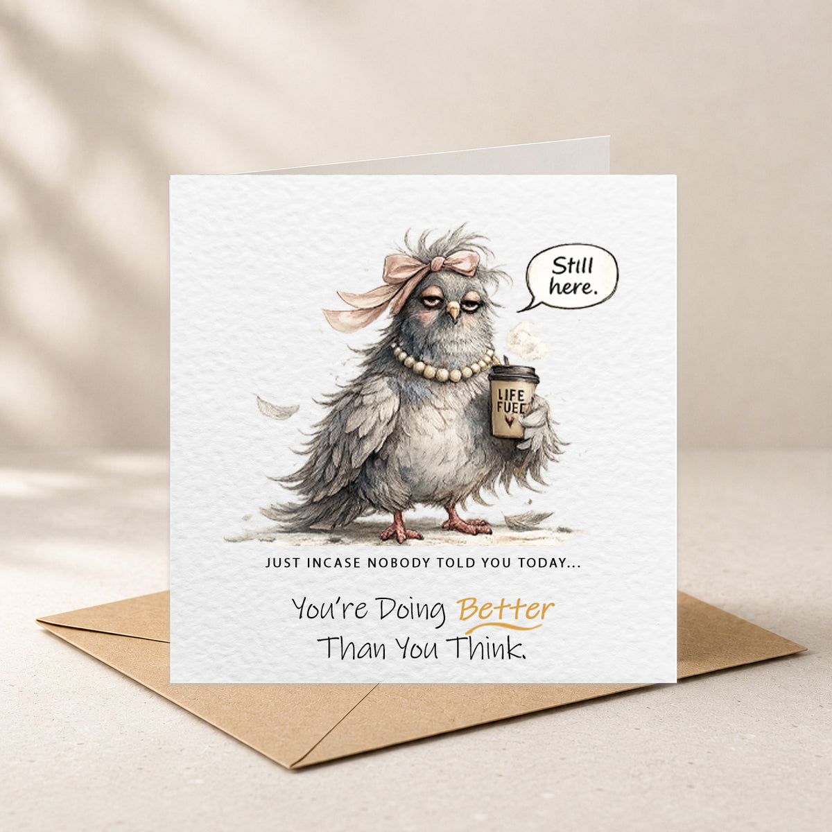 A square greeting card with a brown kraft envelope behind it, featuring an illustrated gray bird wearing a pink bow and pearl necklace, holding a coffee cup and a speech bubble that says 'Still here.' The card reads, 'Just incase nobody told you today.