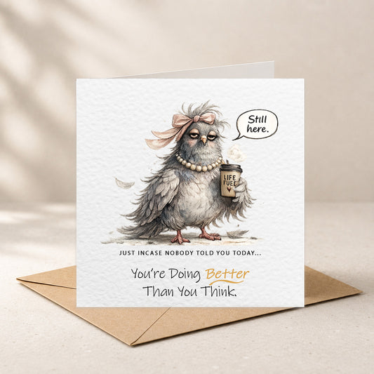 A square greeting card with a brown kraft envelope behind it, featuring an illustrated gray bird wearing a pink bow and pearl necklace, holding a coffee cup and a speech bubble that says 'Still here.' The card reads, 'Just incase nobody told you today.